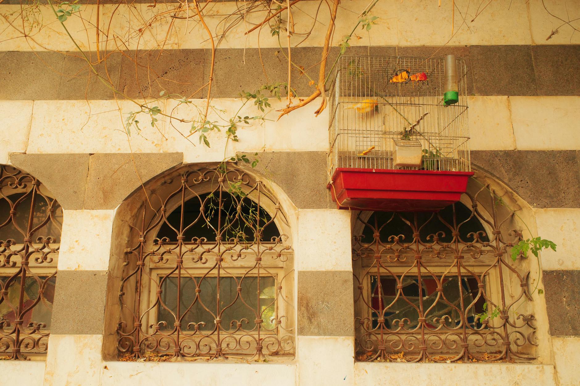 A colorful birdcage hangs above arched windows on a historic patterned facade, evoking timeless charm.