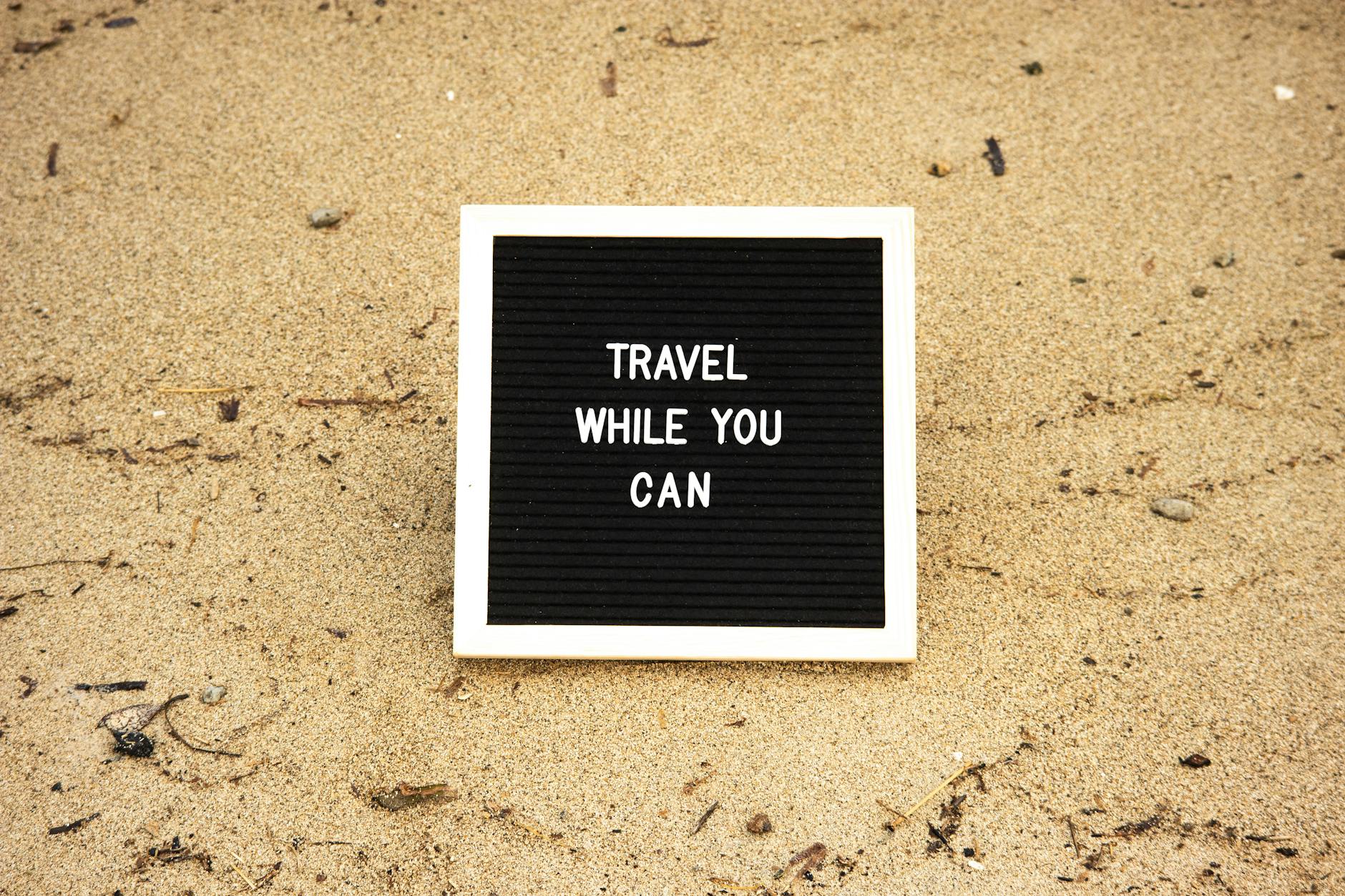 Quote 'Travel While You Can' on board in sand, inspiring travel and adventure.