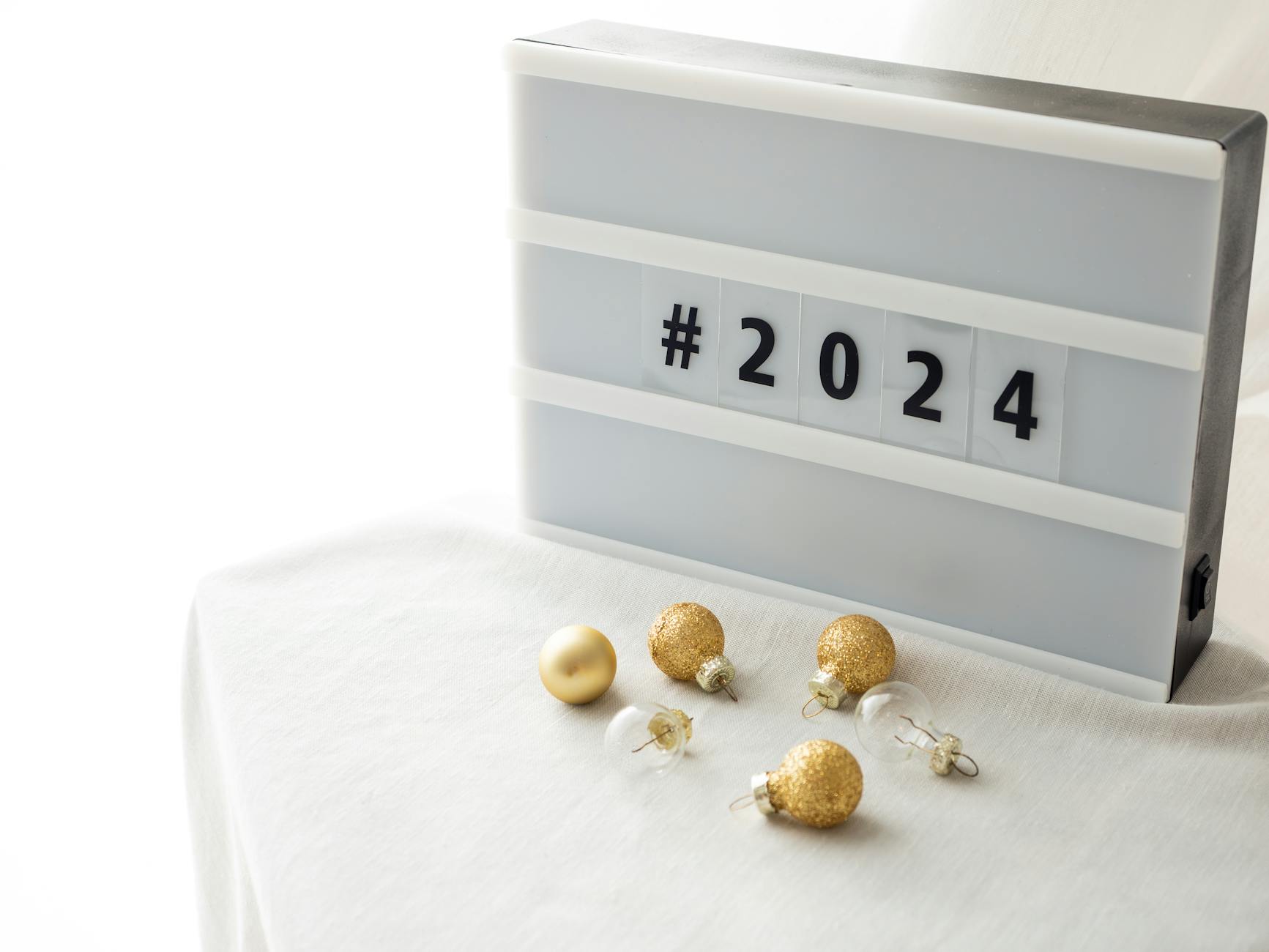 A stylish New Year 2024 decoration with gold baubles and a lightbox on a white tablecloth.