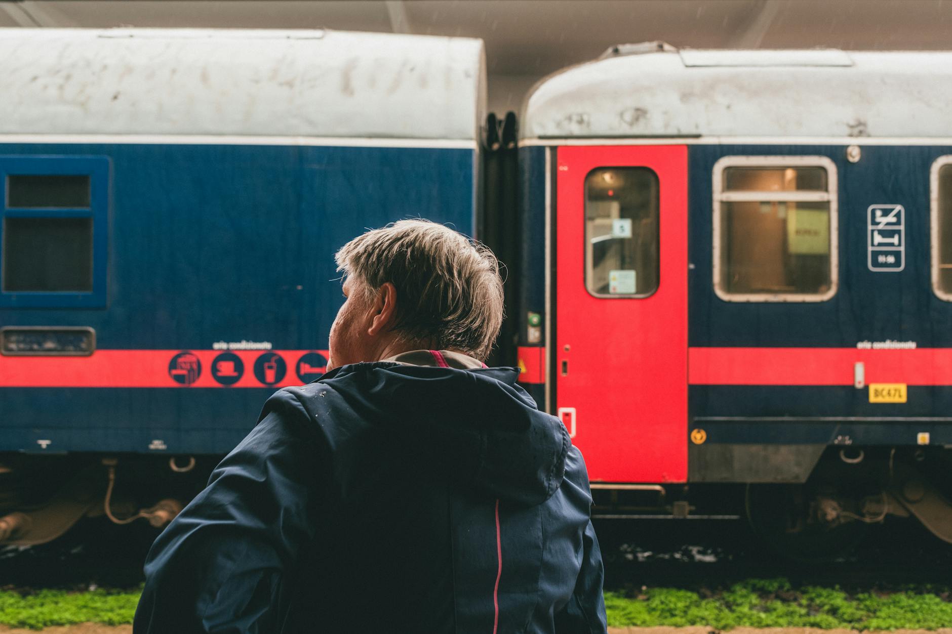 Man waiting by a red and blue train at an outdoor station, viewed from behind.
