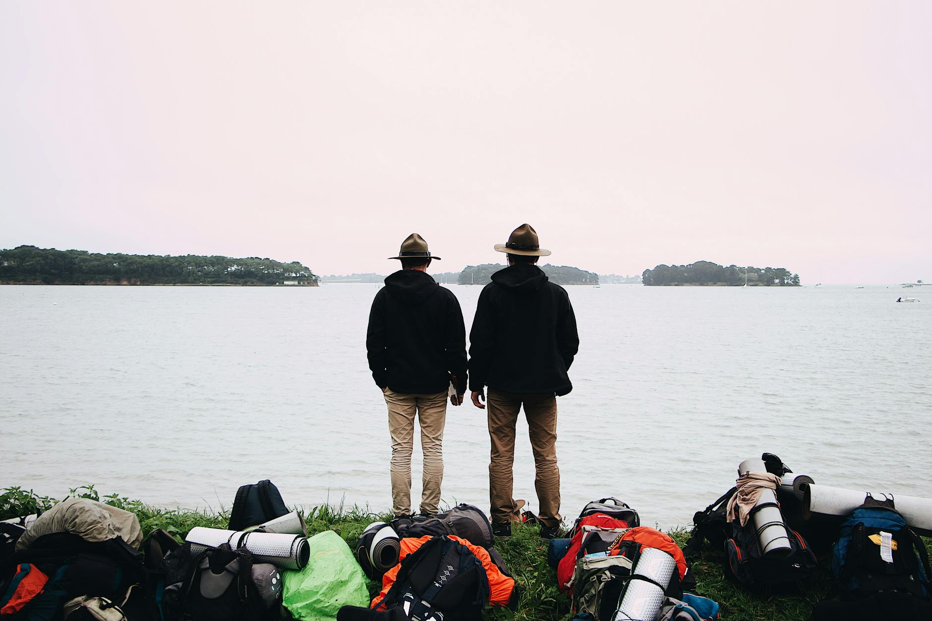 Two scouts in uniform stand by a calm seashore, surrounded by backpacks, ready for an outdoor adventure.