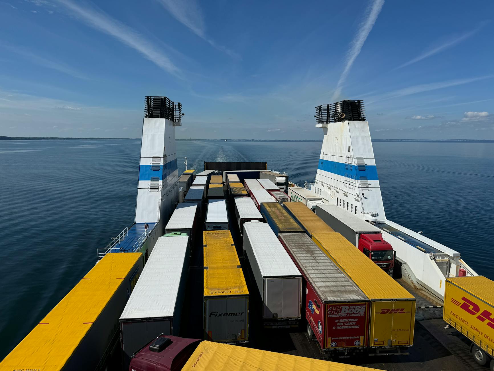 Aerial view of container trucks transported on a large ferry across a calm ocean under a clear blue sky.